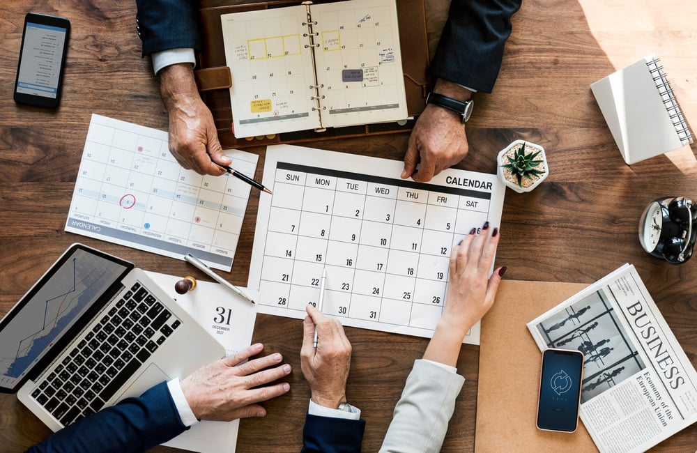 3 business people's hands on a desk with multiple items including calendar, laptop, phones, clock, plant